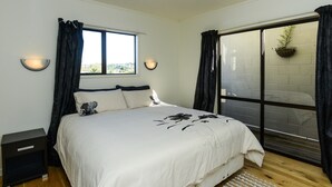 A bedroom with a bed, a nightstand, a window with curtains, a wall-mounted light, and a view through a glass door. - Marine Parade Magic - Napier Holiday Home (Napier City)
