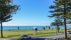 A coastal road with a car driving, a grassy area with benches, and a clear view of the ocean. - Marine Parade Magic - Napier Holiday Home (Napier City)