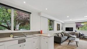 Een moderne keuken met een inbouw eiland, een zithoek met een bank en salontafel, en grote ramen met uitzicht op bomen.