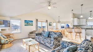 A spacious living room with a large sofa, a coffee table, and a dining area with chairs. There is a kitchen area with cabinets and a window. - Sea Spirit | 717 ft from the beach | Dog Friendly, Private Pool, Hot Tub | Kitty Hawk (Kitty Hawk)