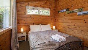 A wooden cabin bedroom with a bed, bedside lamps, and a window. - Nikauscapes - Hot tub in Punakaiki Village (Punakaiki)
