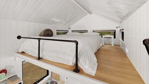 Une chambre à coucher de taille réduite, décorée dans des tons blancs, avec un lit superposé, une petite table de chevet et une fenêtre donnant sur un paysage verdoyant.
