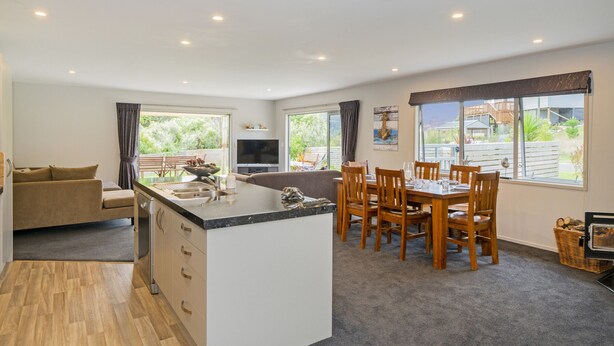A modern kitchen with a central island, dining area with wooden chairs, and large windows offering views of the outdoors. - The Blue Rendevous - Whangamata Holiday Home (Whangamata)