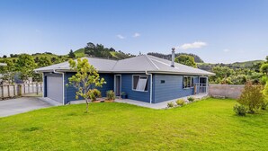 A single-story house with a grey roof and blue siding, a small front yard, and a hill in the background. - The Blue Rendevous - Whangamata Holiday Home (Whangamata)