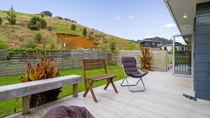 A wooden deck with a bench and a folding chair, overlooking a landscaped garden and a hillside. - The Blue Rendevous - Whangamata Holiday Home (Whangamata)