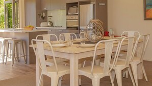 A modern kitchen with a dining area, featuring a wooden table and white chairs, a built-in oven, and a refrigerator. - Cooks Getaway - Cooks Beach Holiday Home (Cooks Beach)