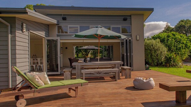 A modern outdoor patio with a wooden deck, a green cushioned lounge chair, a dining table with chairs, and a large umbrella. - Cooks Getaway - Cooks Beach Holiday Home (Cooks Beach)