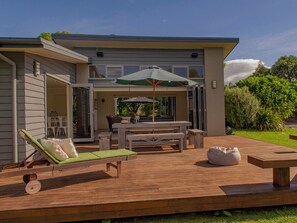 A modern outdoor patio with a wooden deck, a green cushioned lounge chair, a dining table with chairs, and a large umbrella. - Cooks Getaway - Cooks Beach Holiday Home (Cooks Beach)