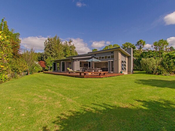 A house with a covered patio, a lawn, and surrounding trees. - Cooks Getaway - Cooks Beach Holiday Home (Cooks Beach)