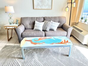 A living room with a grey sofa, two patterned cushions, a colorful coffee table, a small side table with a lamp, and a wall art piece.