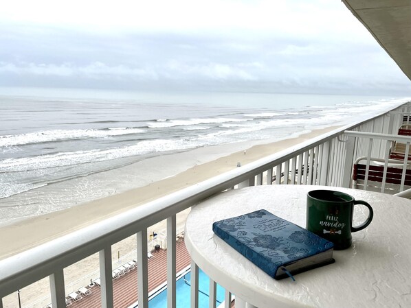 A balcony with a view of the beach and ocean, a blue book, and a green mug.
