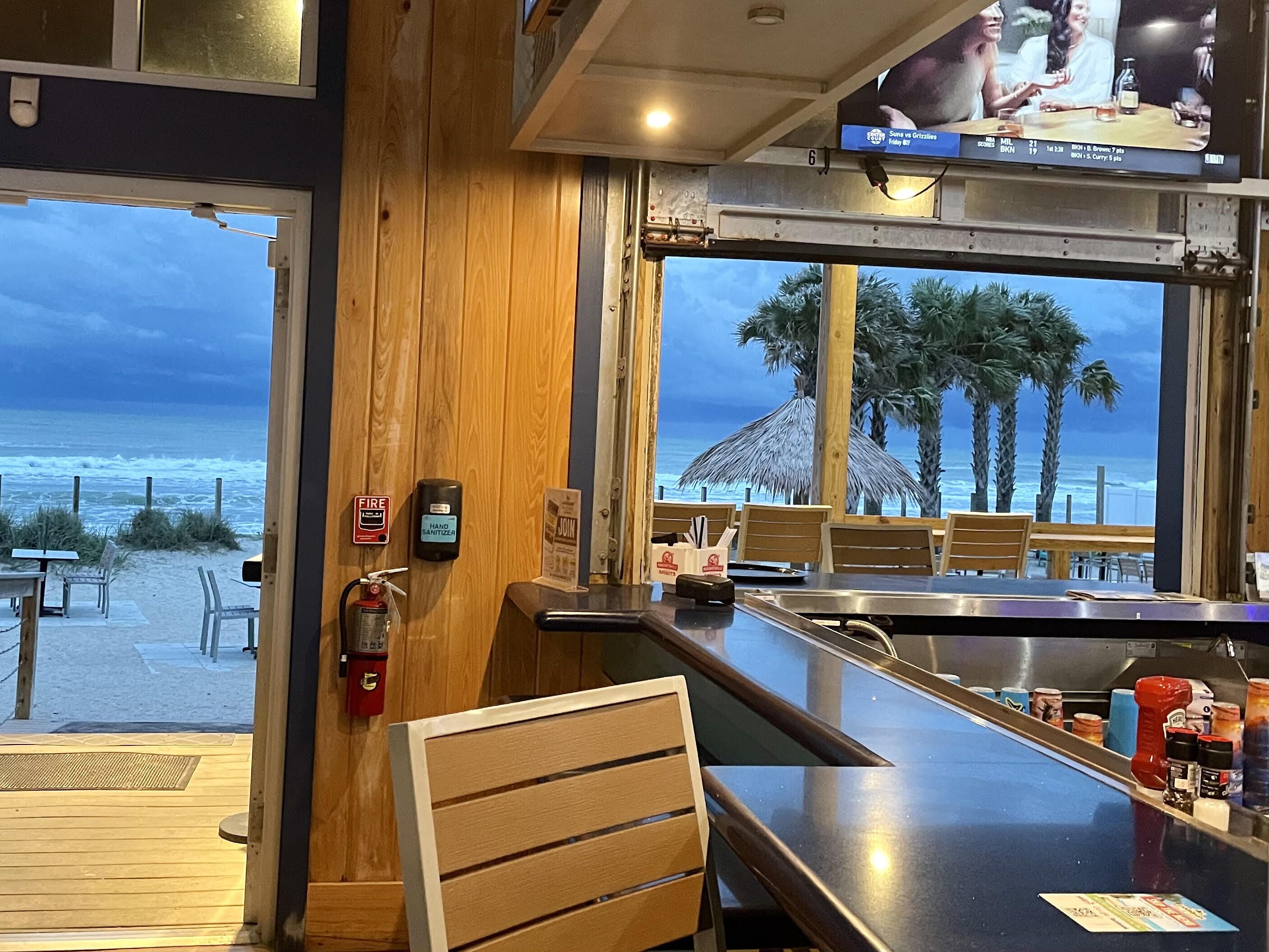 Een bar met uitzicht op het strand en de oceaan, met houten meubels, een brandblusser en een desinfecterende handgeldispenser.