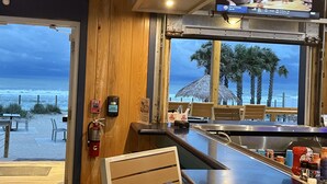 Eine Bar mit Blick auf den Strand und den Ozean, Holzmöbel, ein Feuerlöscher und ein Handdesinfektionsmittelspender.