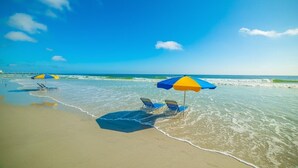Ein Strand mit blauem Himmel, ruhigem Wasser und Liegestühlen unter Sonnenschirmen.