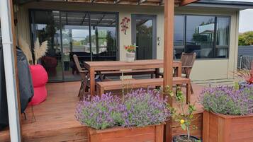 Um deck de madeira com mesa e cadeiras, rodeado por plantas em vasos e uma porta de vidro que dá para uma área interna.