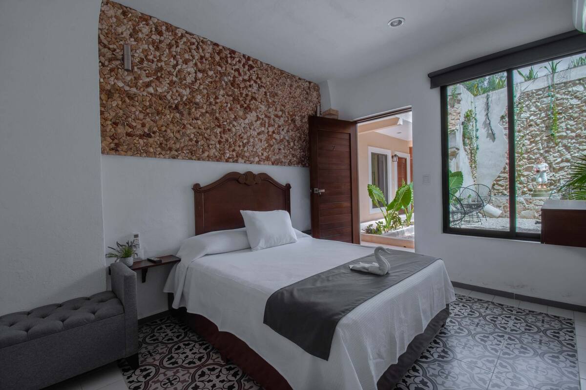 A bedroom with a large bed, a nightstand, a grey sofa, a decorative wall, and a view of an outdoor area with plants and a statue.