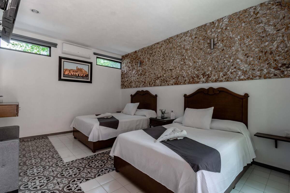 A hotel room with two beds, a patterned rug, a wall-mounted air conditioner, and a framed picture on the wall.