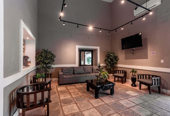 A modern living room with a sofa, coffee table, and chairs, a flat-screen TV mounted on the wall, and ceiling lights.