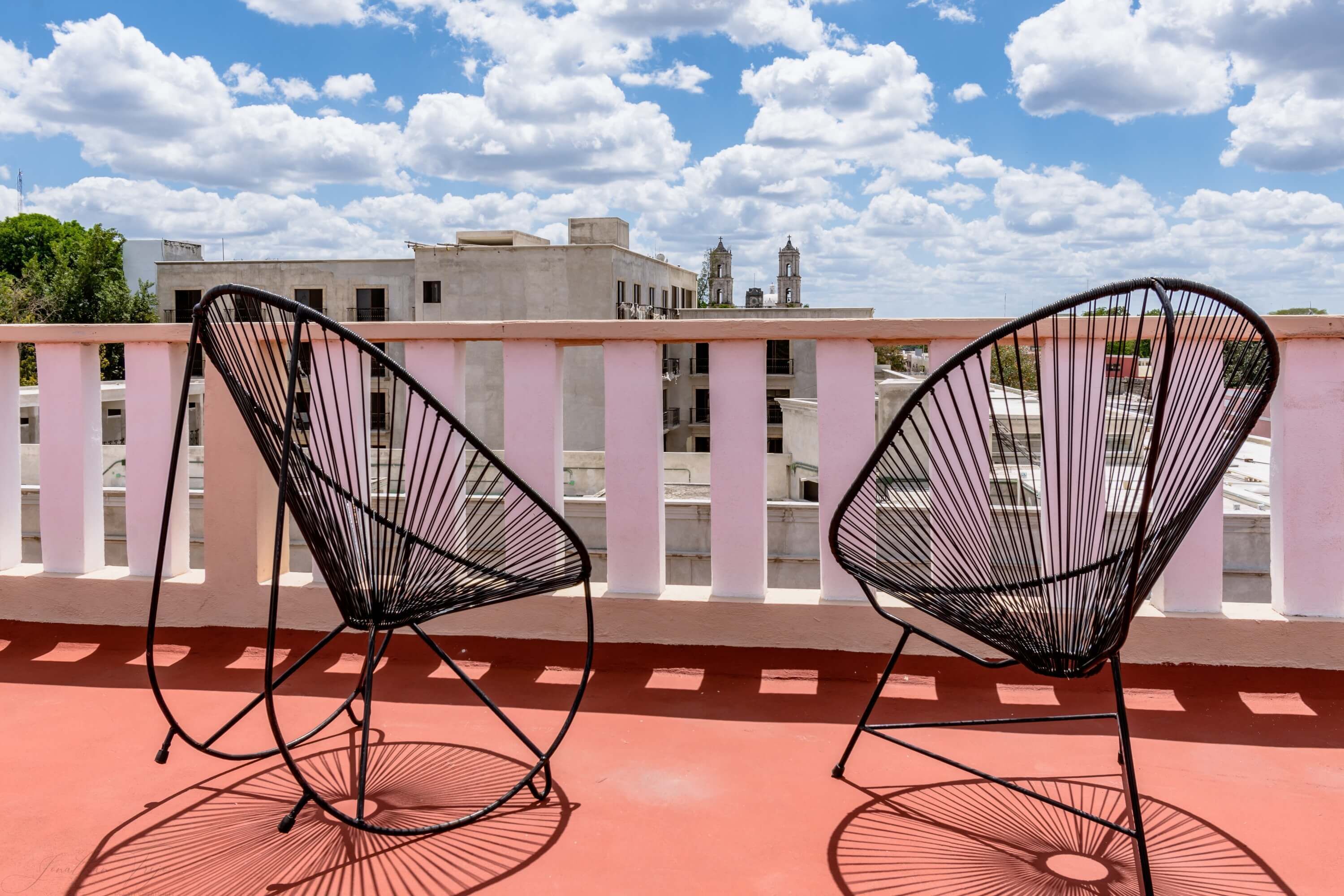 Two black wireframe chairs on a red-tiled balcony with a white railing, overlooking a cityscape with buildings and a clear sky.