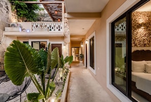A hotel corridor with a covered outdoor seating area, large plants, and a balcony with a railing.