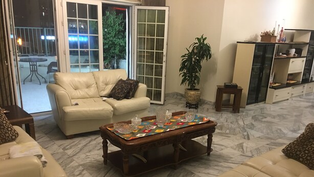 A living room with a beige sofa, a coffee table with decorative items, a potted plant, and a kitchen area in the background.