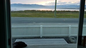 Uitzicht door een deur op een terras met een witte picket fence, een hond liggend op het terras, en een waterpartij met bergen op de achtergrond.