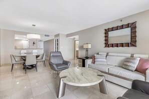 Una sala moderna con una mesa de centro redonda de mármol, un sillón de cuero gris, un sofá blanco con cojines estampados y una zona de comedor con una mesa y sillas de vidrio.
