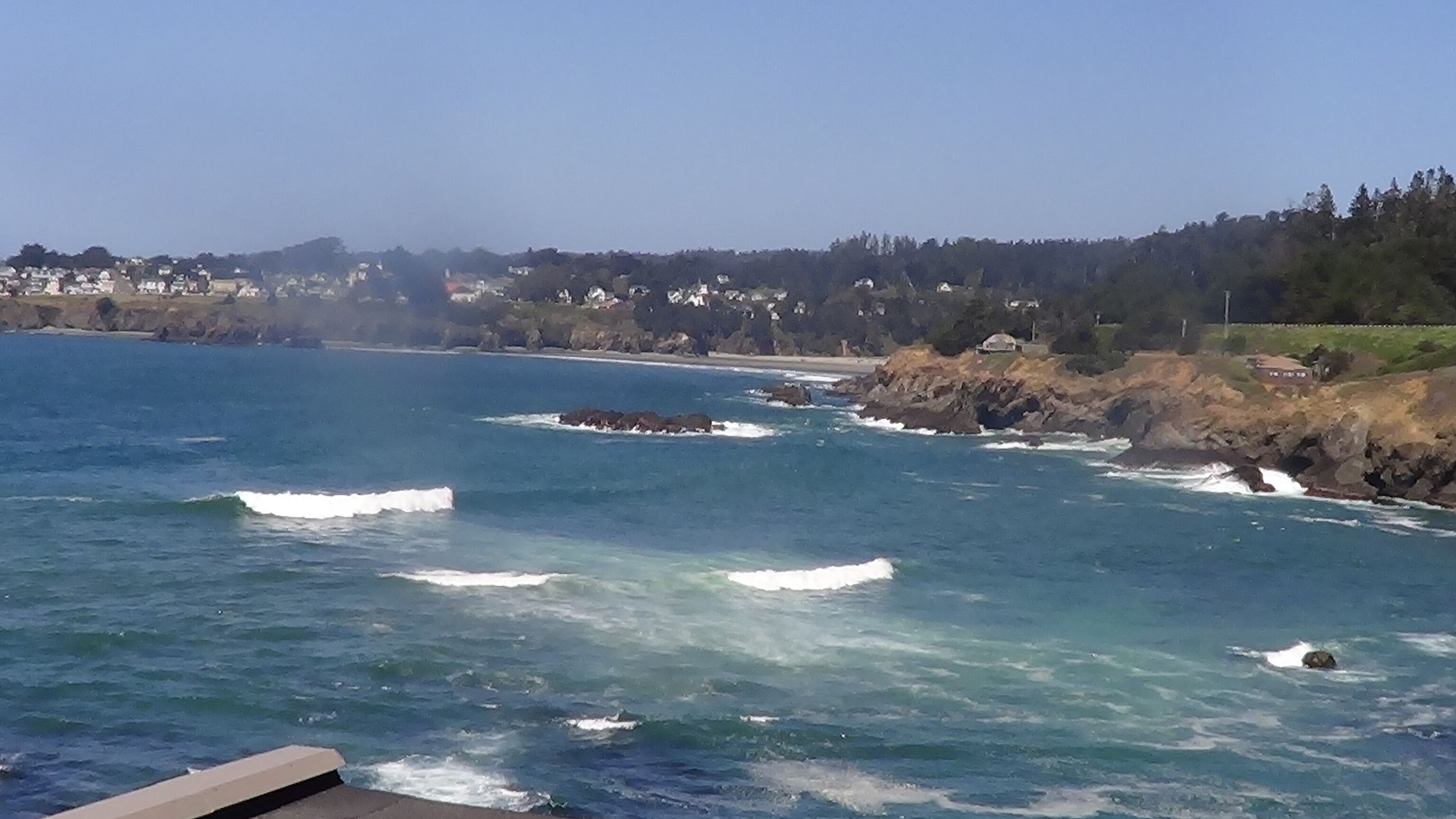 A coastal view with waves crashing against rocks, a clear sky, and a distant shoreline with buildings.