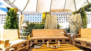 Cómodo espacio exterior con un sofá acolchado y sillas de madera, una mesita con un libro y vasos, y una carpa encima.