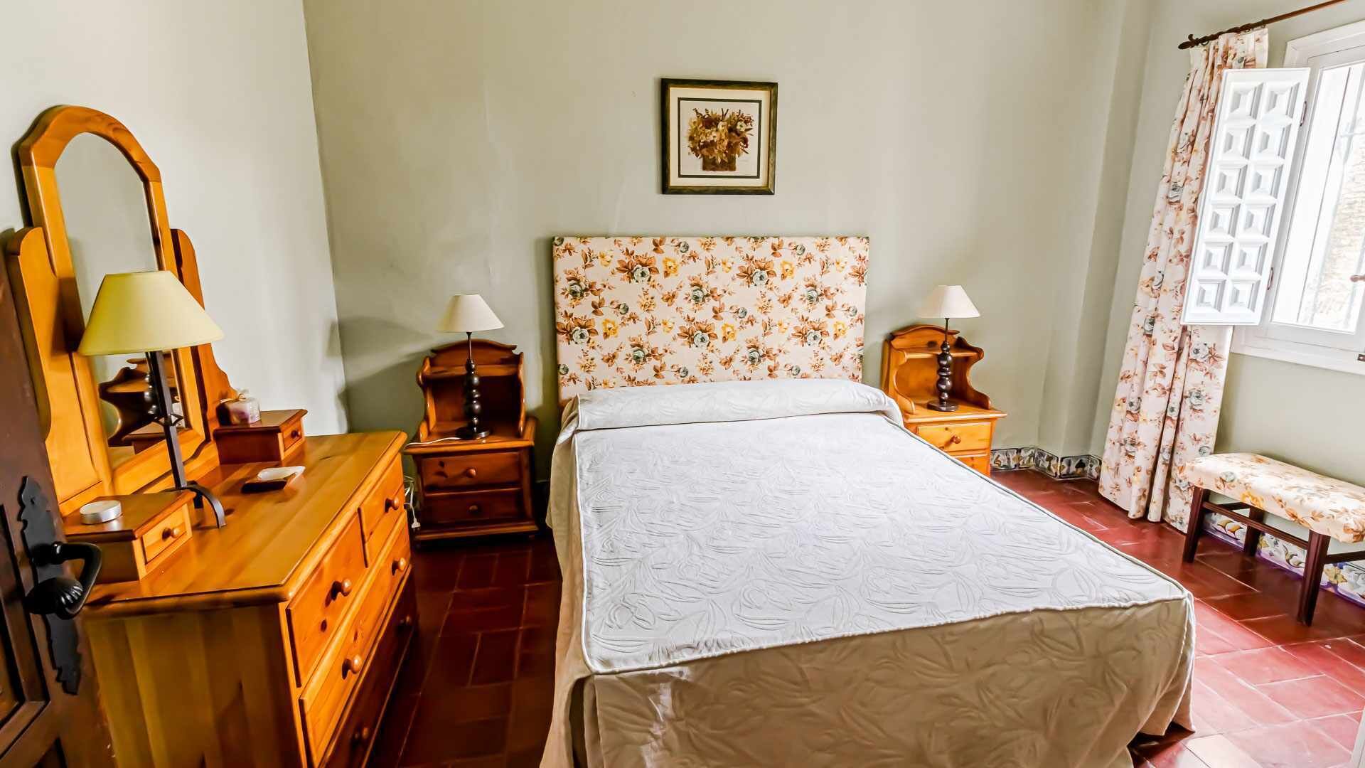 Quarto com cama, cômoda de madeira, mesas de cabeceira, luminárias, painel de cabeceira floral, quadro na parede e janela com cortinas.