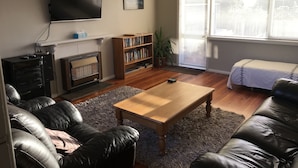 Ein Wohnzimmer mit Kamin, Wand-Fernseher, Bücherregal, Couchtisch und einem Fenster mit Jalousien.