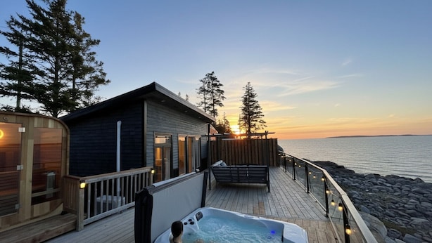 Una terraza de madera con un jacuzzi, una cabaña pequeña y vistas al atardecer sobre el agua.