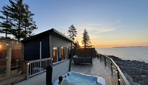 A wooden deck with a hot tub, a small cabin, and a view of the sunset over the water.