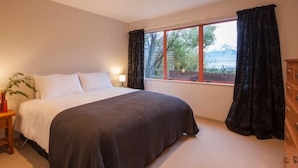 Un dormitorio con una cama grande, una ventana con cortinas, una mesita de noche con lámpara y un aparador.