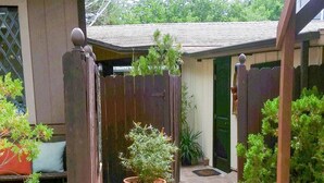 Un étroit sentier mène à une maison en bois dotée d’un porche, qui est entourée de plantes en pot et d’une clôture en bois.