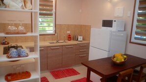 Een compacte keuken met een spoelbak, magnetron en koelkast. Er staat een eettafel met een fruitschaal en een rood vloerkleed.