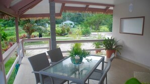 Een overdekt terras met een glazen tafel en rieten stoelen, omringd door potplanten en uitzicht op een weelderige tuin en een huis op de achtergrond.