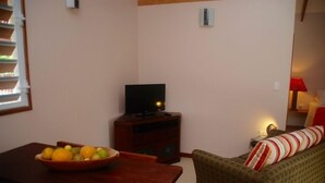 Een gezellige woonkamer met een houten tafel, een televisie op een standaard, een bank met een gestreept kussen en een raam met jaloezieën.