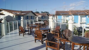 Een terras met houten stoelen en tafels, een witte balustrade en gebouwen met betegelde daken op de achtergrond.