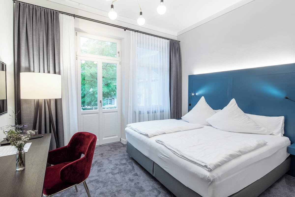 Habitación de hotel con cama, escritorio con lámpara, una silla y ventana con cortinas.