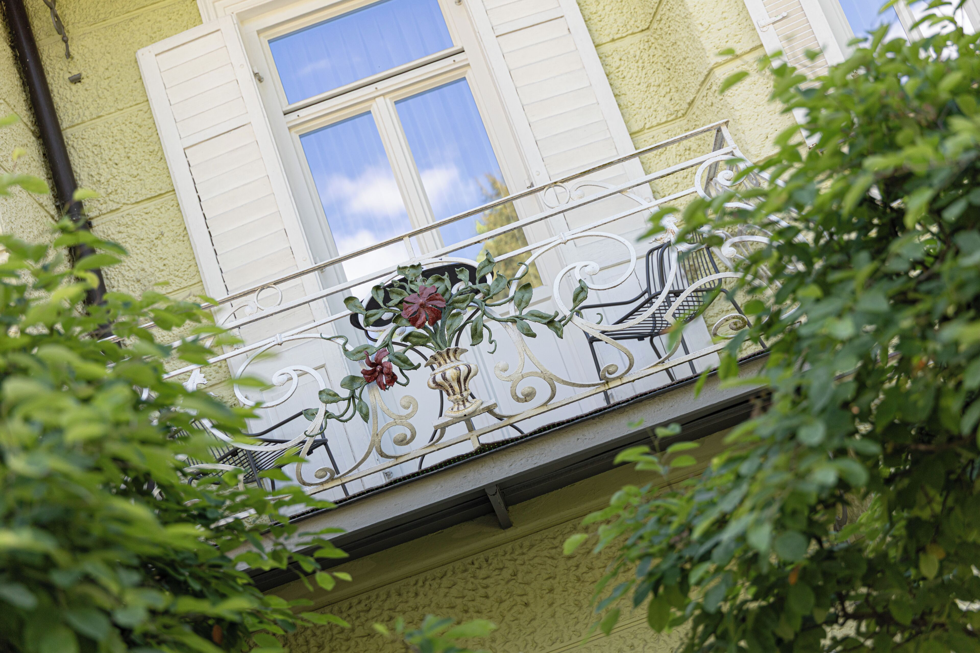 A balcony with a wrought iron railing, a potted plant, and a window with shutters.