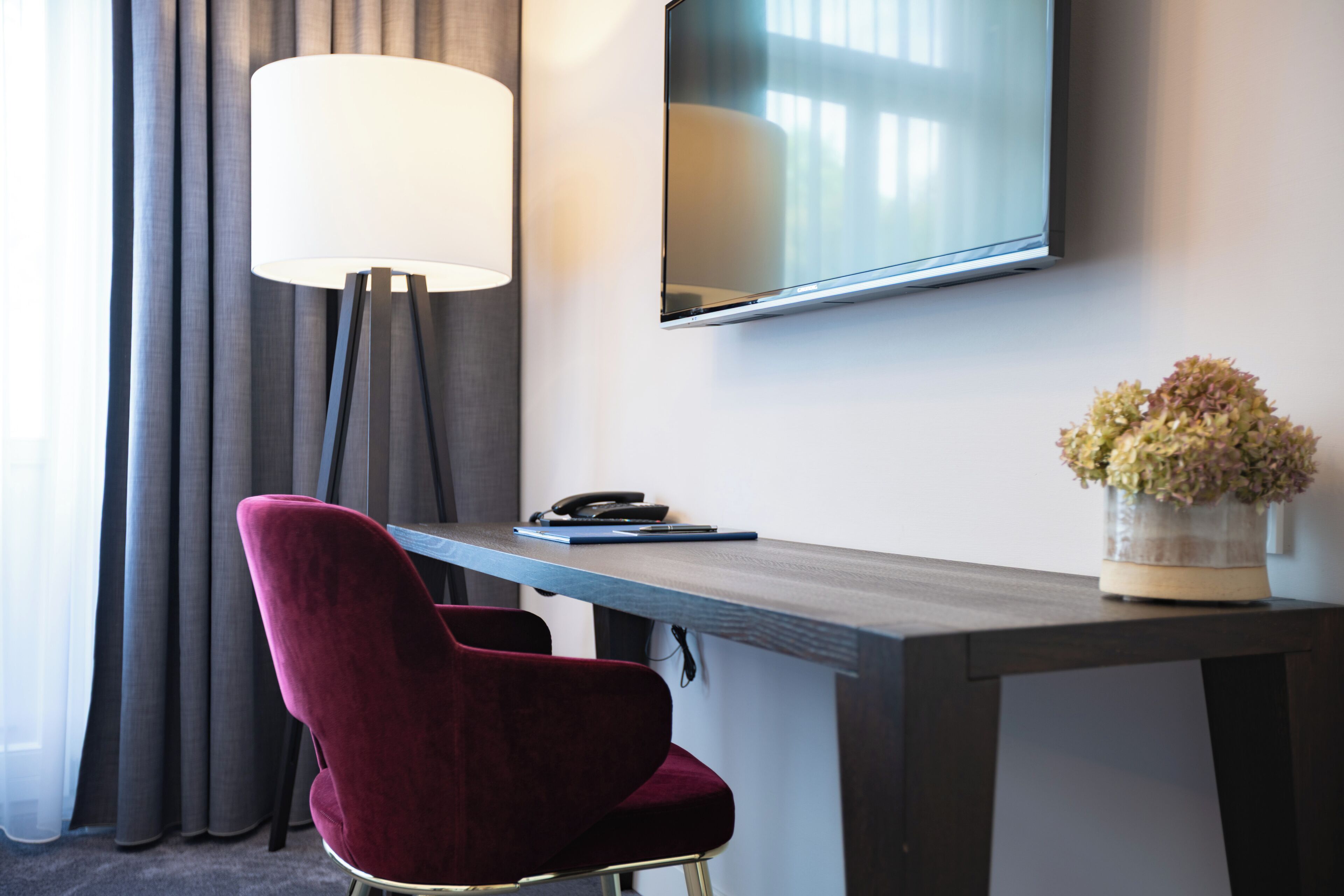 Een hotelkamer met een bureau, een rode stoel, een lamp, een flatscreen televisie en een vaas met bloemen.