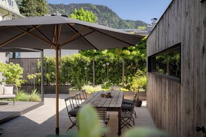 Patio esterno con tavolo in legno, sedie e un ombrellone grande.