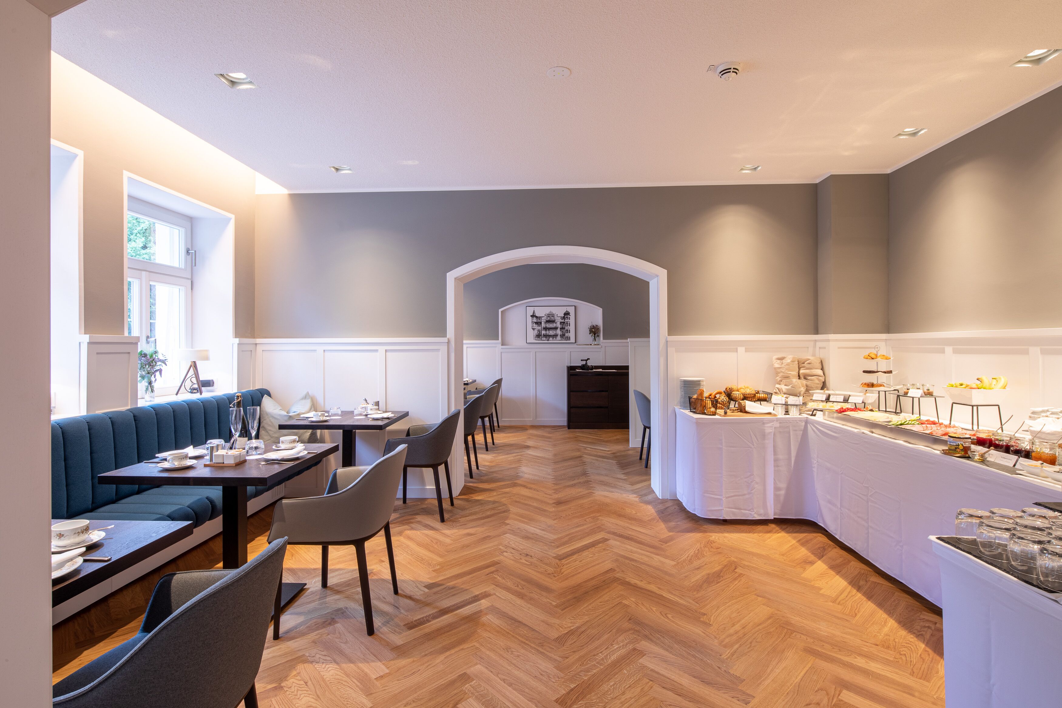 Un comedor moderno con piso de madera en patrón espiga, una mesa larga para compartir y una zona de estar con un sofá azul.