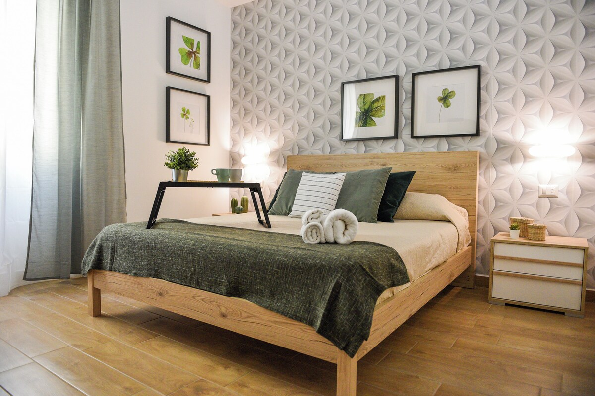 Ein ordentlich bezogenes Bett mit grüner Decke, ein Nachttisch mit Lampe und eine Wand mit geometrischen Mustern.