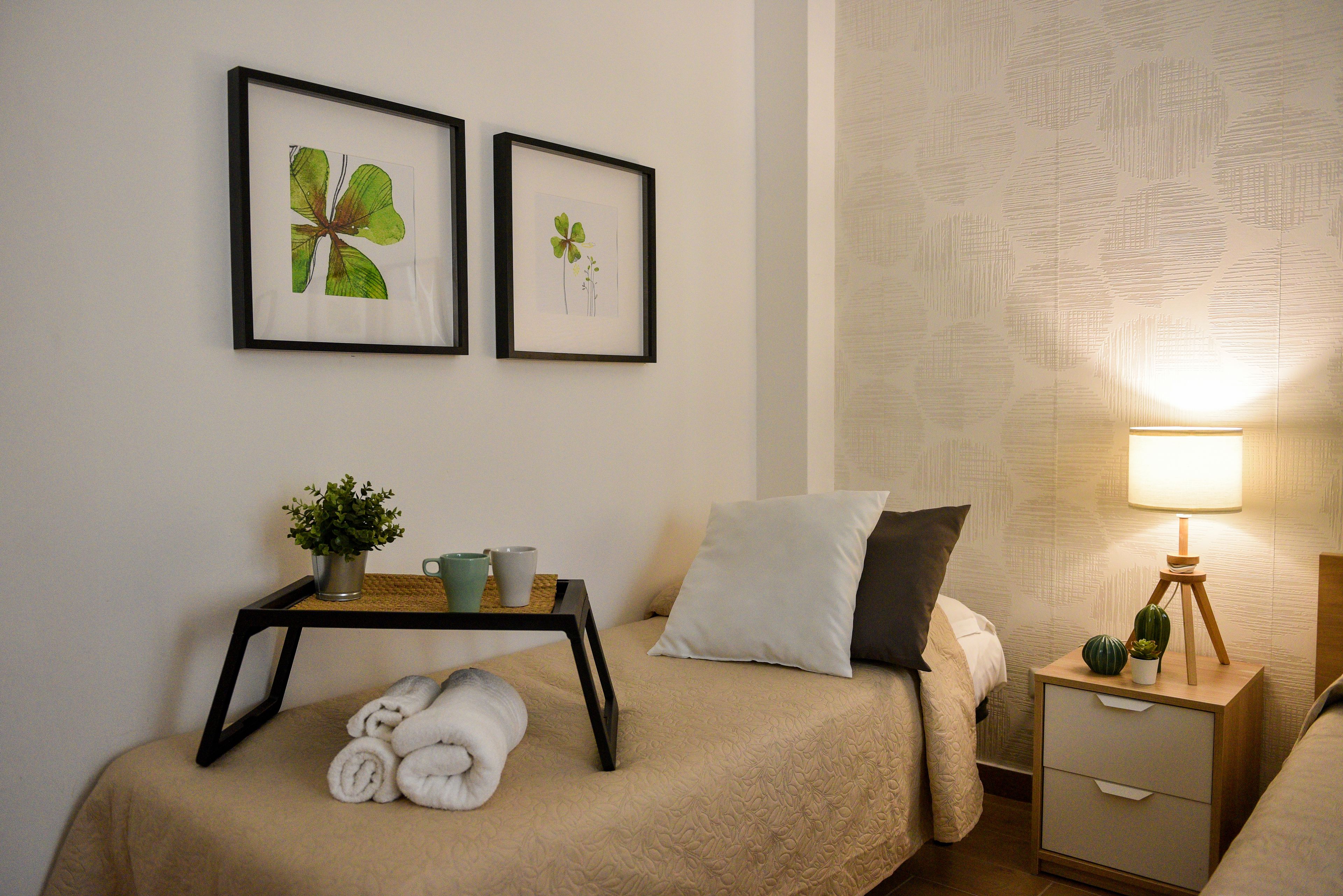 Un cuarto de hotel ordenado con una cama, una mesita de noche con una lámpara, una planta pequeña e ilustraciones botánicas enmarcadas en la pared.