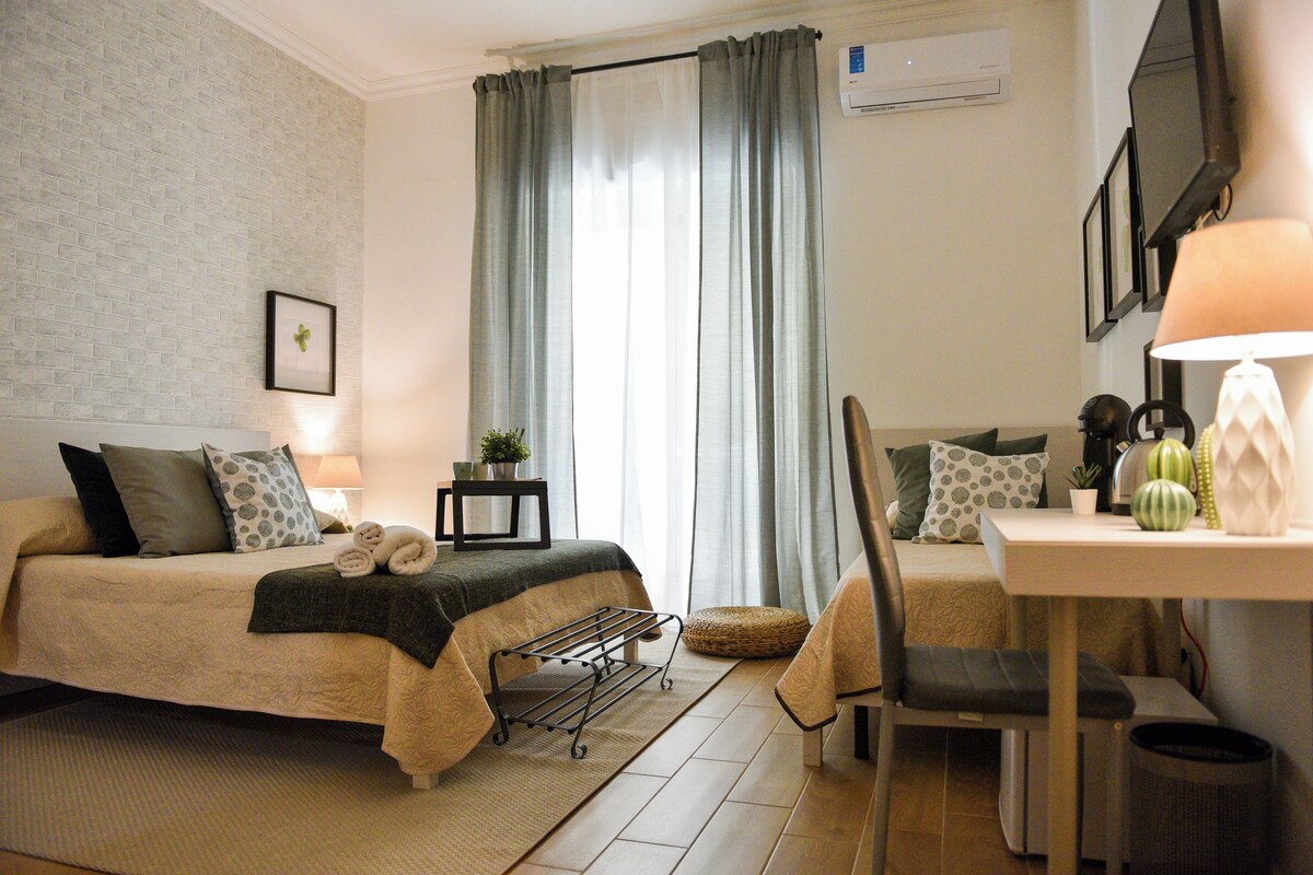 Ein Hotelzimmer mit einem Bett, einem Schreibtisch, einem Sessel, einem kleinen Tisch, einem Mülleimer, einer Zimmerpflanze und einem Fenster mit Vorhängen.