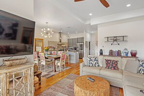 A spacious living area with a sofa, coffee table, and a TV. There is a dining area with chairs and a table, and a kitchen in the background.