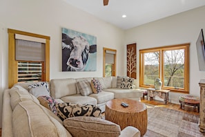 A living room with a sectional sofa, a round wooden coffee table, a large painting of a cow, and a window with a view of trees.
