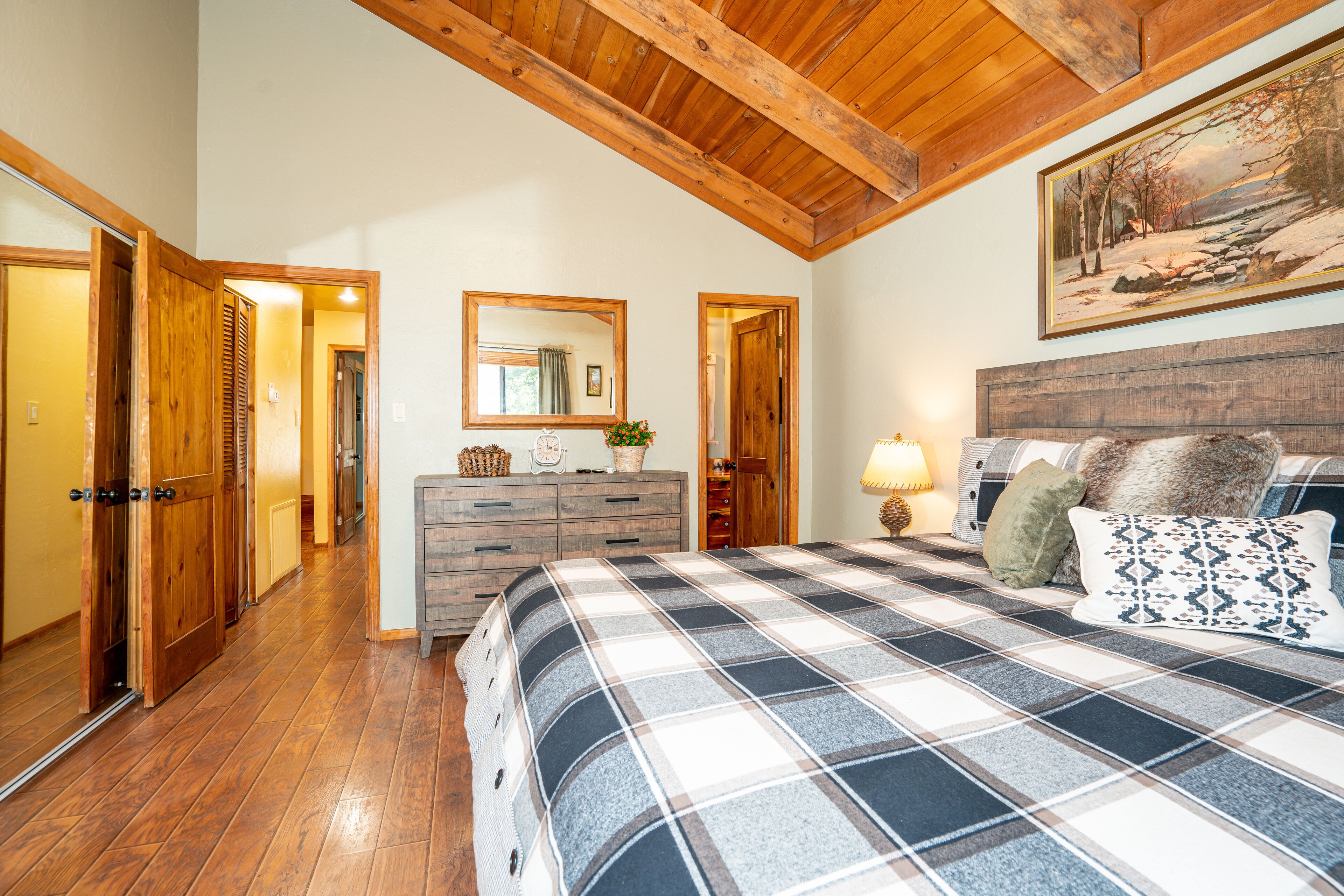 A bedroom with a plaid bedspread, wooden ceiling beams, a dresser, a mirror, and a painting on the wall.